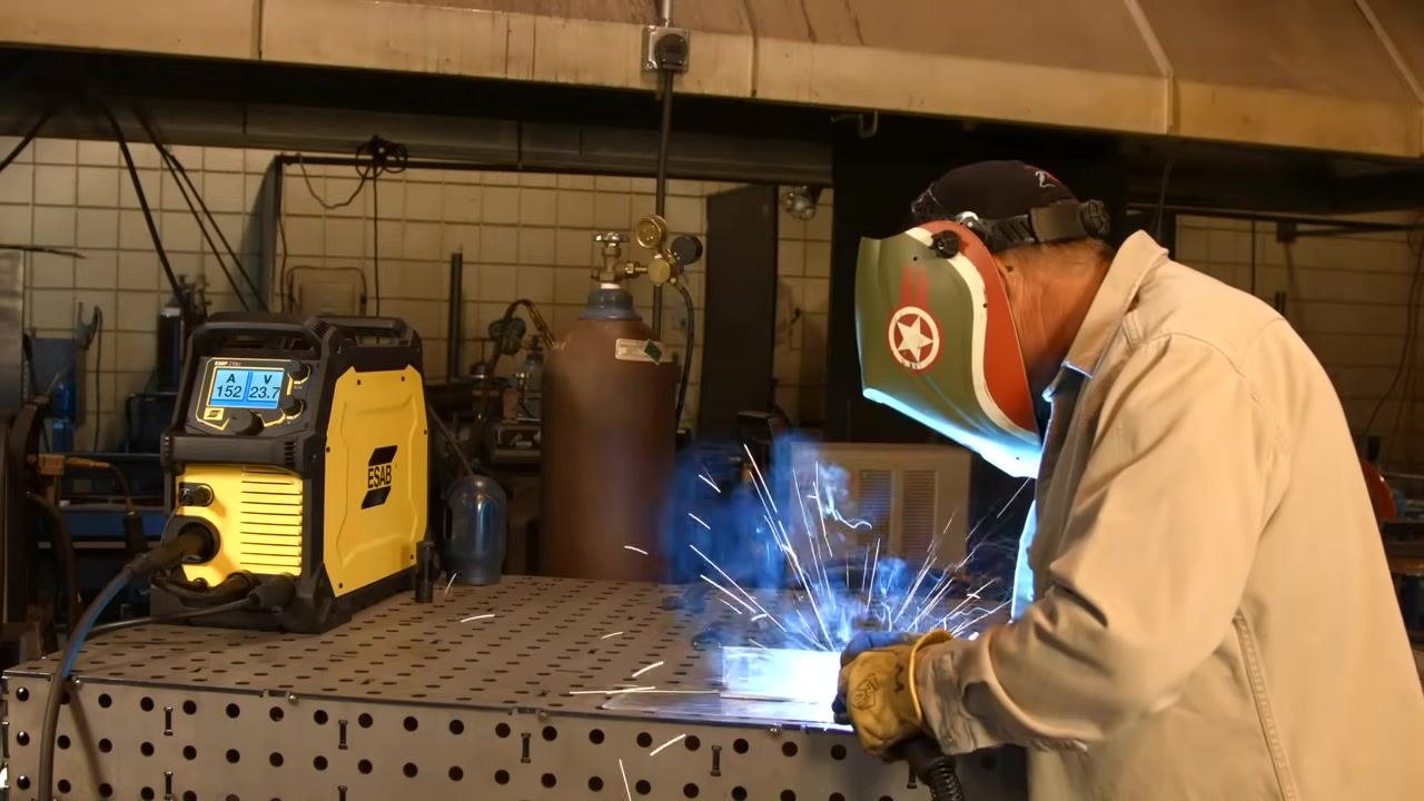 Active welding with visible arc and sparks; the welding machine display shows live amperage and voltage.