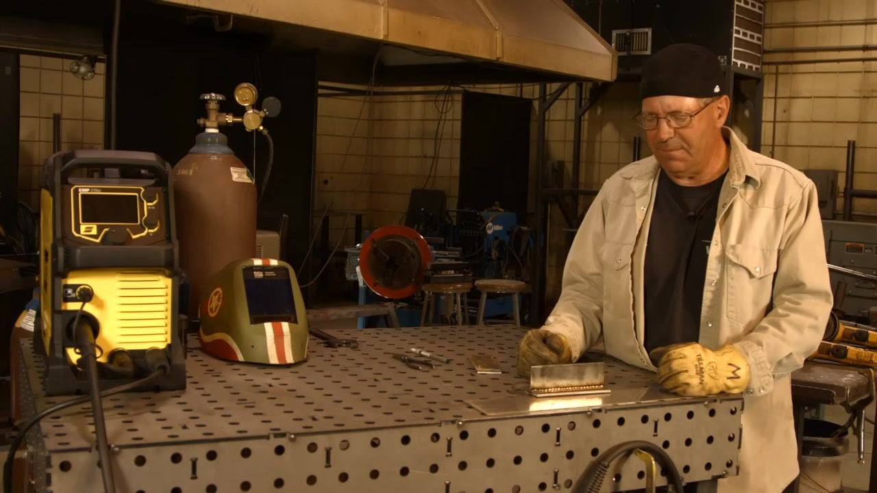 Operator standing at the welding table with the machine, gas cylinder, and helmet, reviewing weld results and machine features.