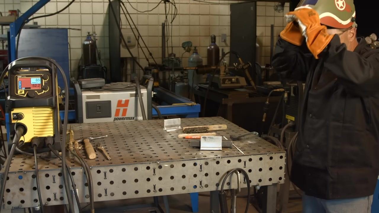Putting on a welding helmet at the workbench, with the ESAB EMP 210 welder and various tools visible, ready for welding.
