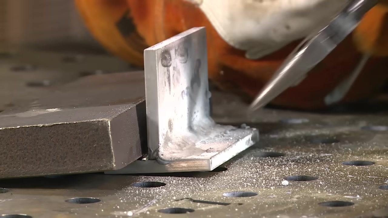 Close-up of the welded joint showing visible slag and spatter, with a gloved hand holding a tool near the weld and debris on the table surface.