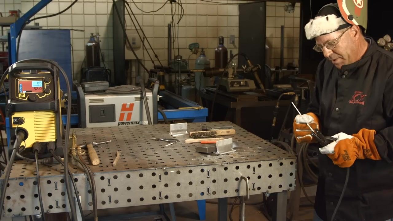 Preparing to weld with electrode and electrode holder in hand, the welding machine set to 85A/52.0V, and tools and metal pieces on the table.