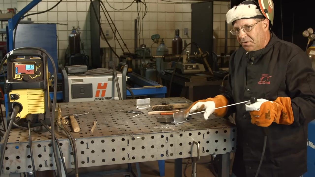 Gesturing toward the welded joint with the electrode while discussing grinding and multiple passes, with the welding machine and tools visible.