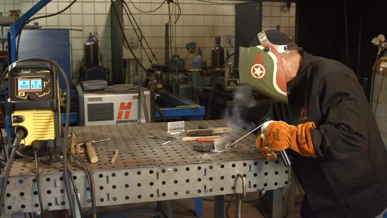 Actively welding with the helmet down and the arc visible; the welding machine display shows 101A/17.5V, with tools and materials on the table.