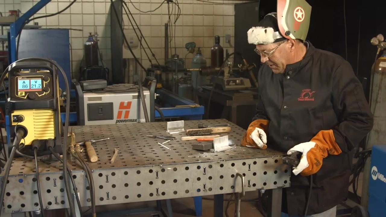 Helmet raised while inspecting the welded joint; the machine display shows 86A/21.2V, with tools and materials on the table.