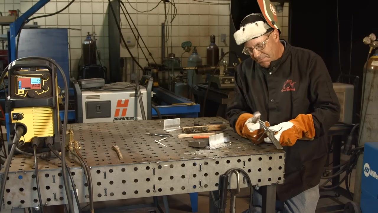 Using a tool to lightly tap and remove slag from the welded joint, with gloves and helmet on and the welding table's tools and materials visible.