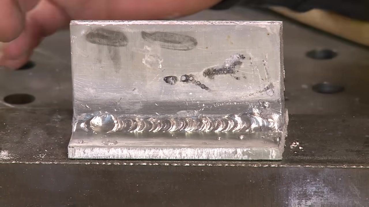 Close-up of an aluminum weld joint showing spatter, weld bead, and workshop setting.
