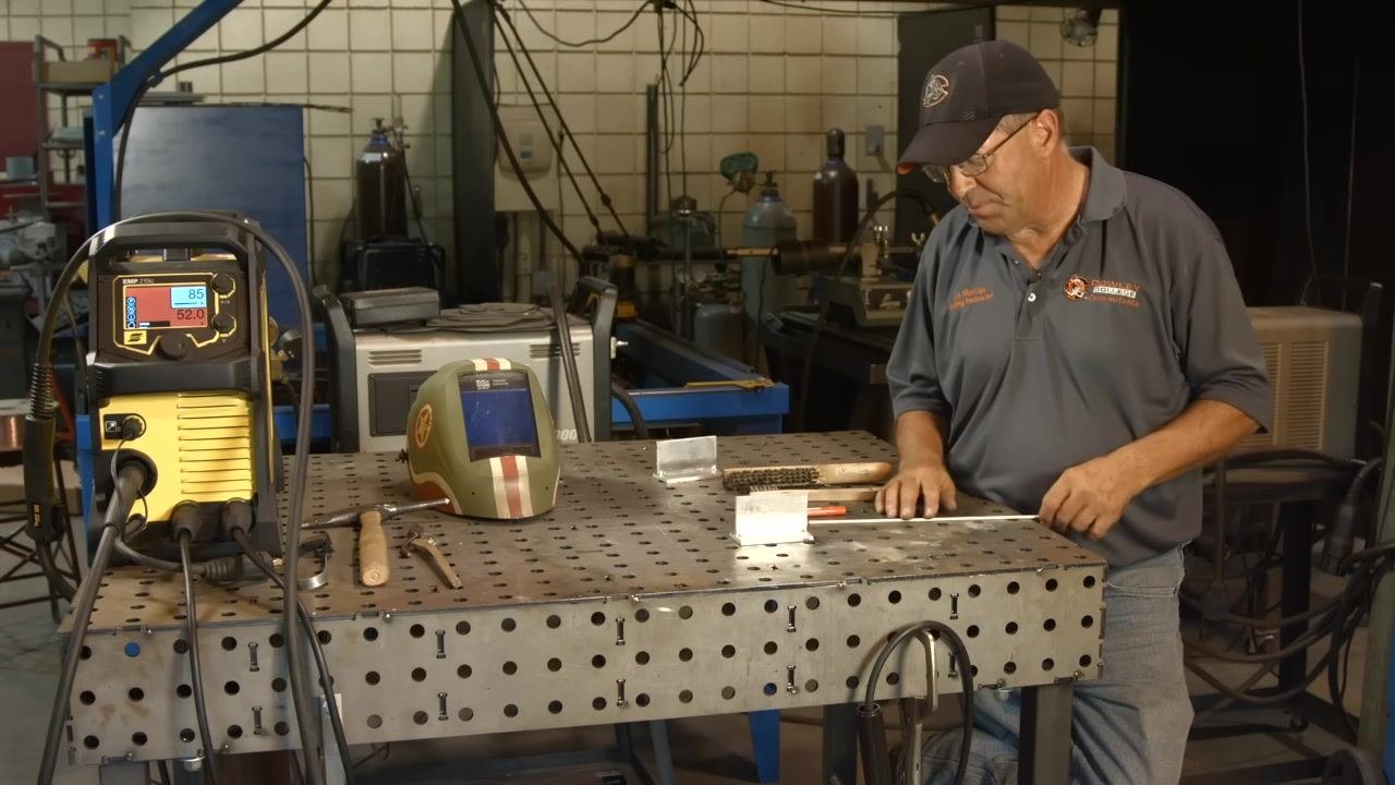 A welding table holds an ESAB EMP 210 welder displaying 85 amps and 52.0 volts, along with a welding helmet, wire brush, marker, and aluminum workpieces staged for the preheating process.