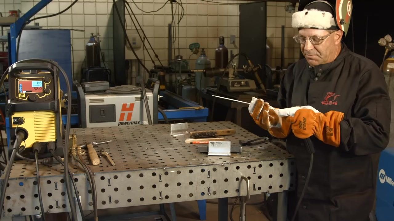 Full PPE in place while holding a low hydrogen electrode and electrode holder; the welding machine is set to 85A/52.0V, with tools and metal pieces on the perforated welding table.