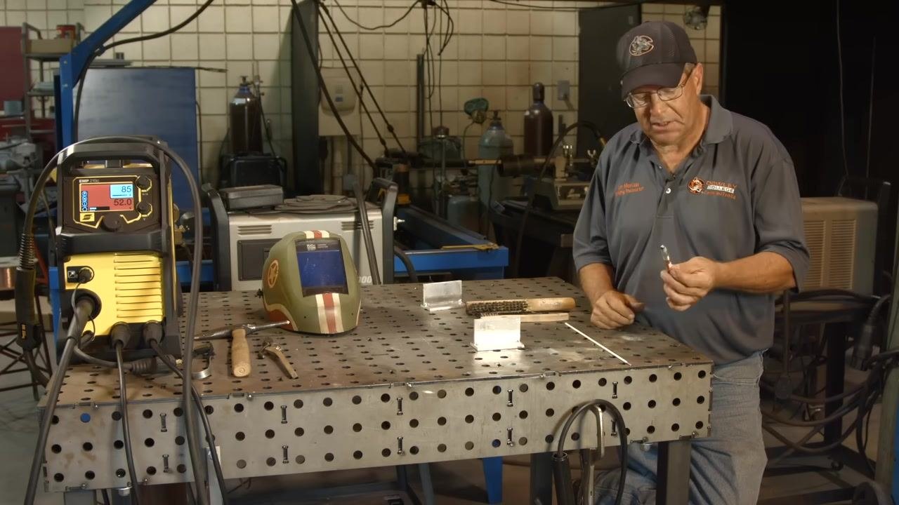 Holding a temperature-indicating stick (Tempilstik) in preparation to mark the aluminum workpiece, with a welding helmet, wire brush, and aluminum pieces visible on the workbench.
