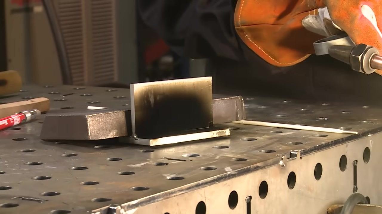 Applying a neutral flame from an oxy-acetylene torch to the aluminum workpiece, which shows a visible soot layer on the welding table.