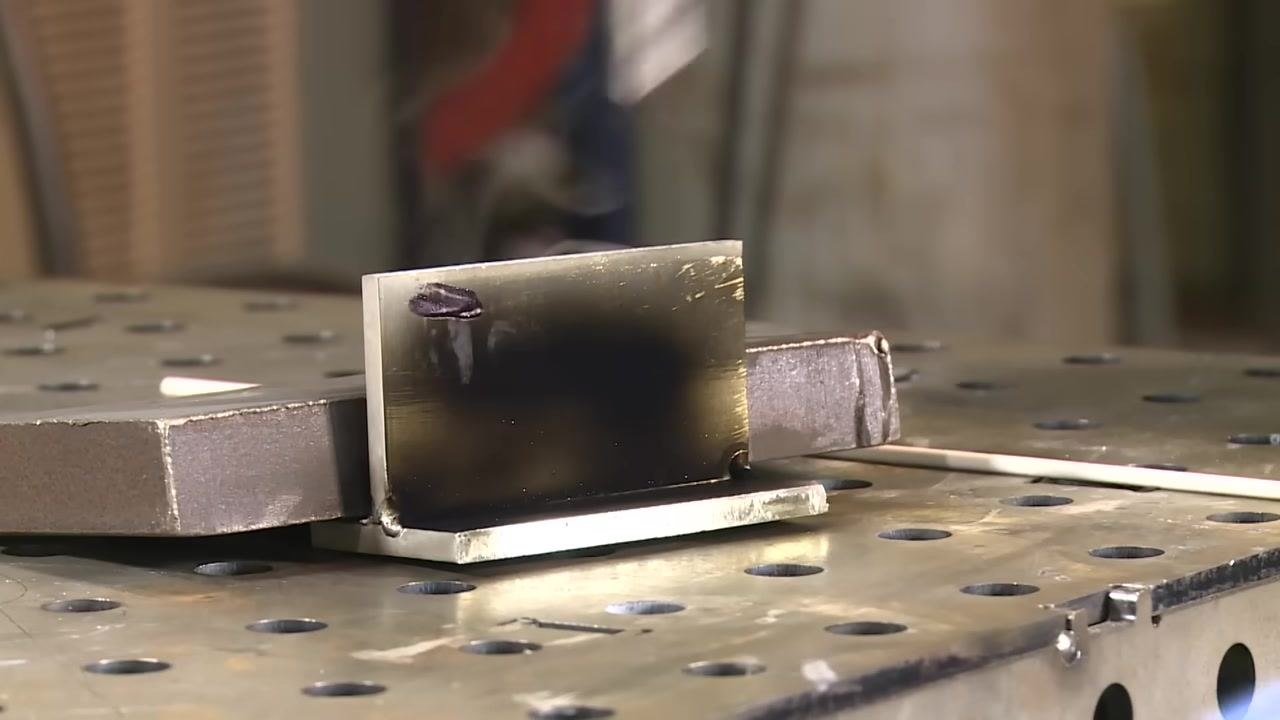 An oxy-acetylene torch flame applied to the aluminum workpiece, now cleaner with visible tack welds, sitting on a perforated welding table.