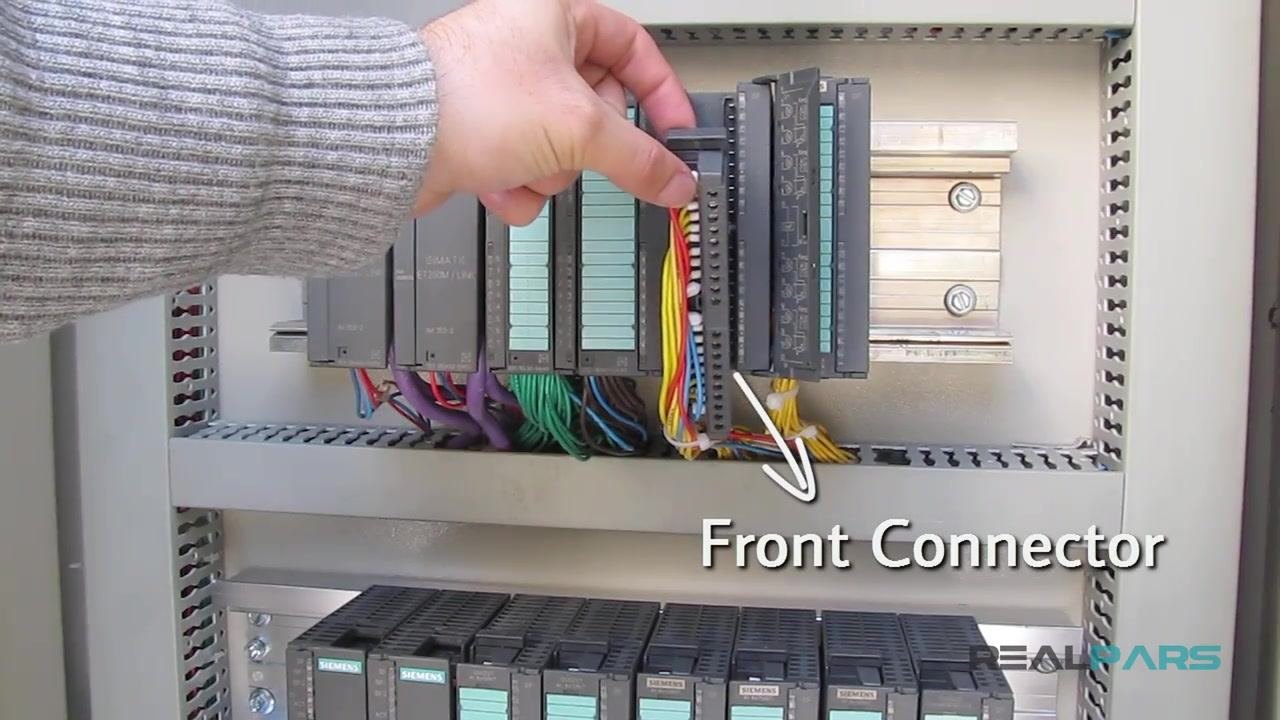 A hand installing a front connector onto a Siemens PLC module inside a control cabinet, with multiple colored wires (yellow, red, blue) attached. Other PLC modules and organized wiring are visible.