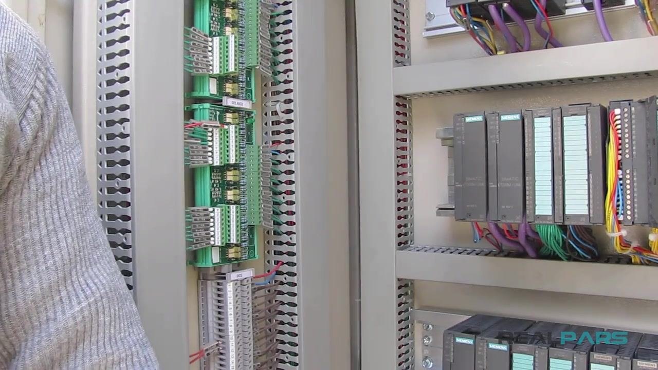 A wire stripper removing insulation from the end of a red wire, with the PLC cabinet and terminal blocks visible in the background.