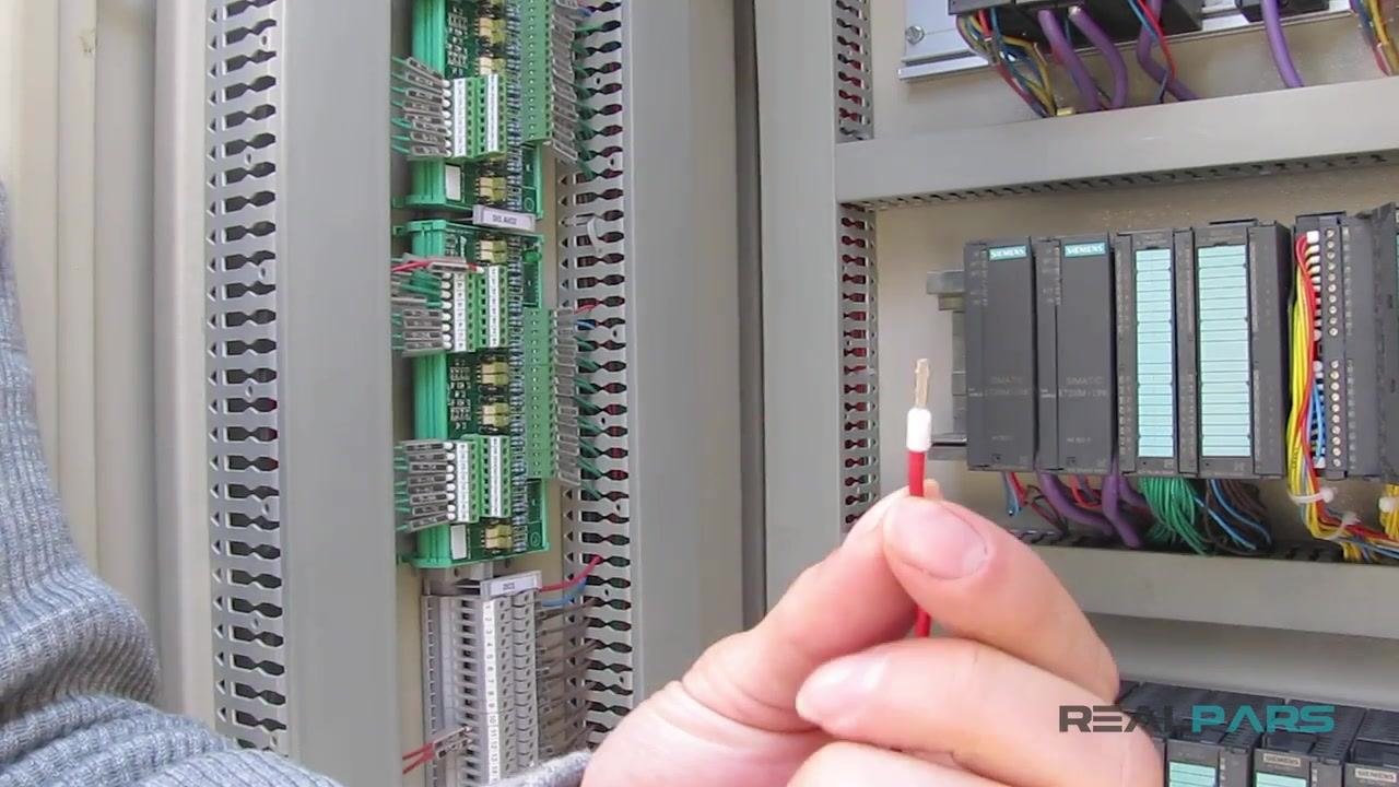 A red wire with a stripped end and a white ferrule being crimped with a yellow-handled crimping tool, with the PLC control cabinet visible in the background.