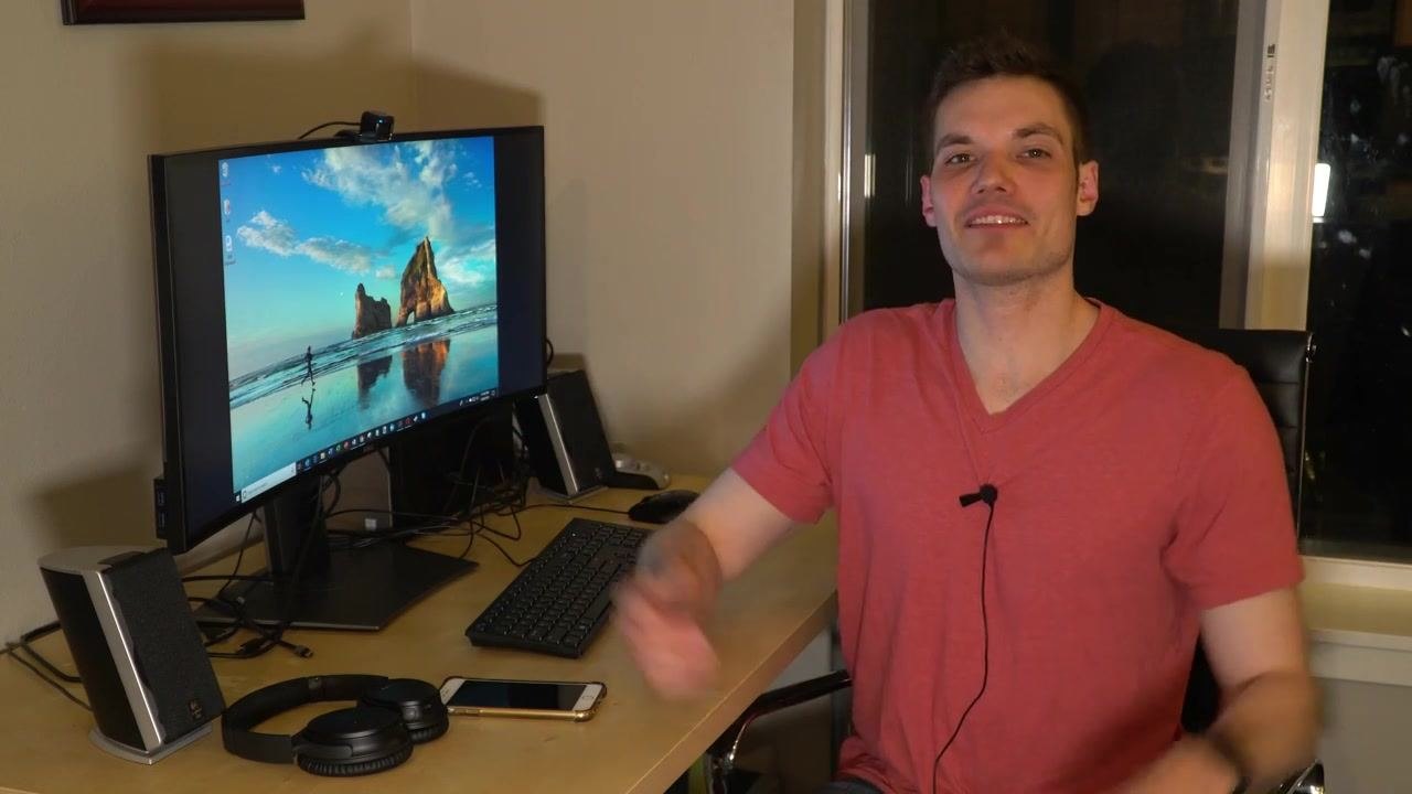 A person sitting at a desk with a monitor displaying the Windows desktop, along with a keyboard, mouse, headphones, and a smartphone in a home office setting.