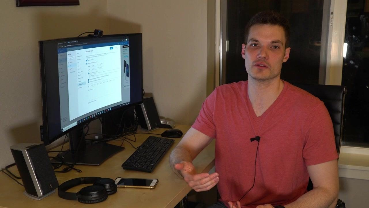 A person sits at a desk in a home office environment, facing the camera and waving with a smile. On the desk are a curved monitor displaying the Outlook web app's Automatic Replies settings, a keyboard, mouse, headphones, smartphone, and speakers. The room is warmly lit, and the presenter appears to be wrapping up the video.