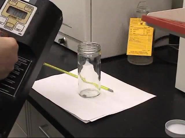 A laboratory worker sits on a stool while an EH&S staff member holds a mercury vapor detector nearby, in a lab with cabinets and an emergency shower.