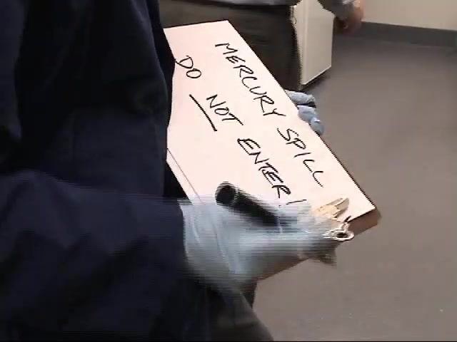 A worker in a lab coat and gloves writes "MERCURY SPILL DO NOT ENTER" on a clipboard to create a warning sign.