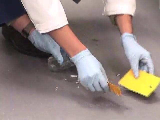 A gloved worker uses two pieces of paper to scoop up mercury beads and broken glass, placing them into a glass jar.