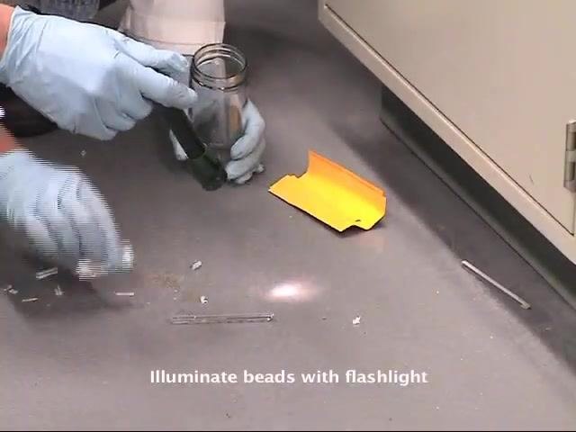 A gloved worker uses a flashlight and glass jar to inspect the floor near a cabinet for remaining mercury beads and broken glass.
