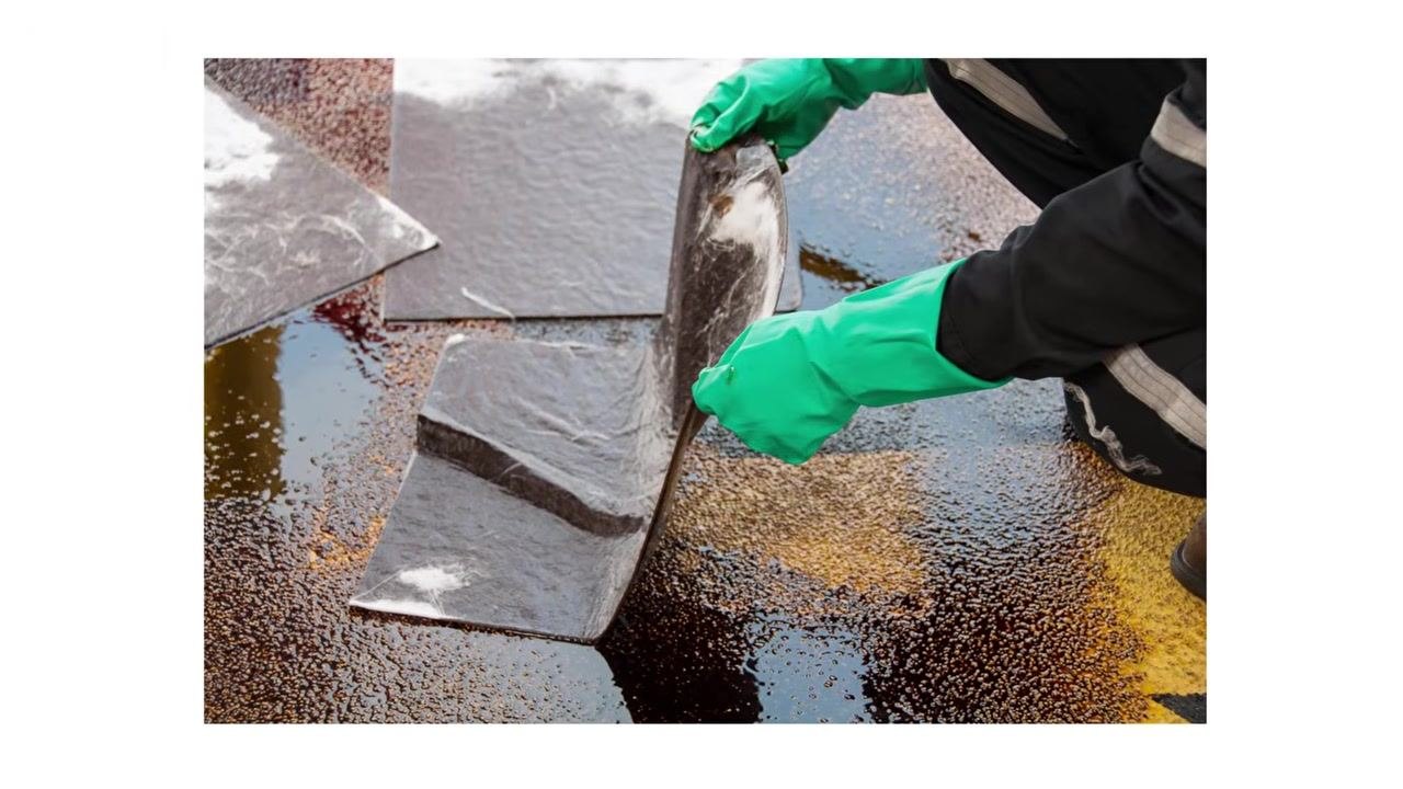 Person wearing green chemical-resistant gloves placing absorbent pads onto a liquid chemical spill on the ground in an industrial or outdoor setting, with visible liquid and multiple absorbent pads in use