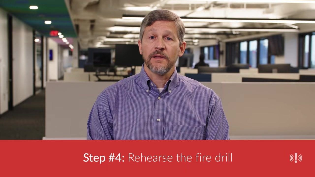 A person in a purple checkered shirt seated in front of open-plan cubicles, with a banner reading "Step #4: Rehearse the fire drill."