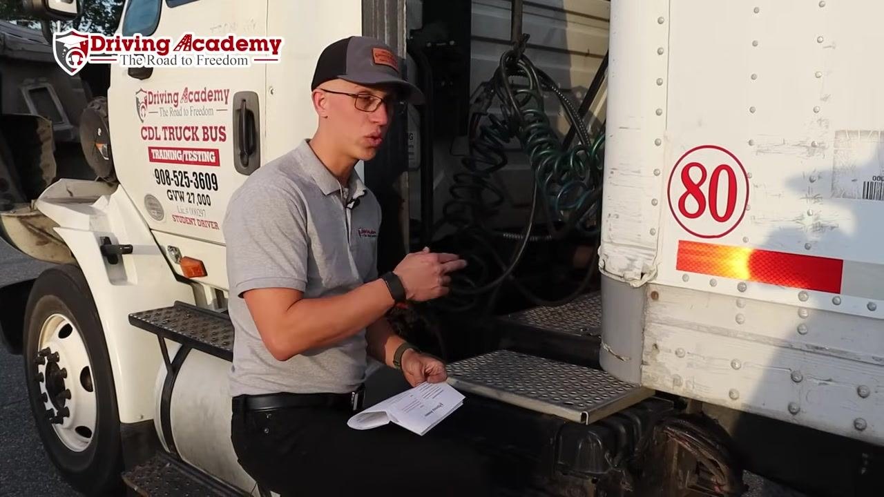 Instructor sitting on the truck step, holding a checklist and preparing to discuss the frame between tractor and trailer.