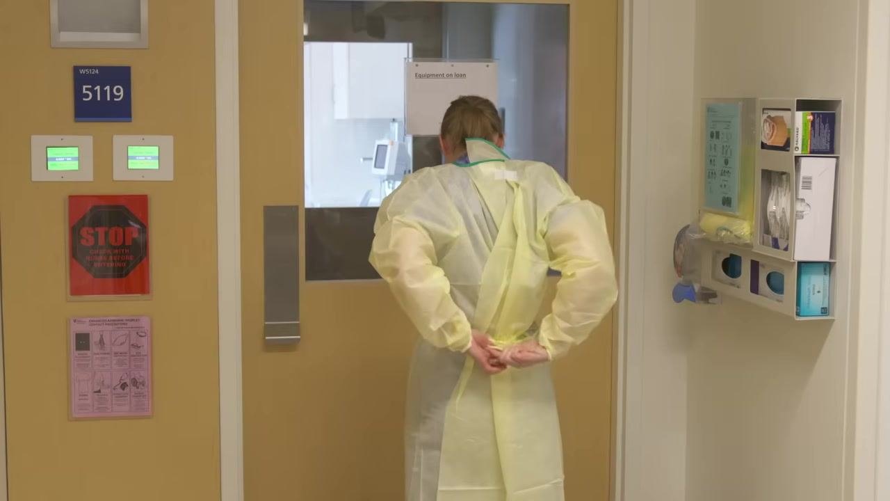 Healthcare worker placing the bottom strap of an N95 respirator over the back of her neck, wearing a yellow gown, with hair tied back.
