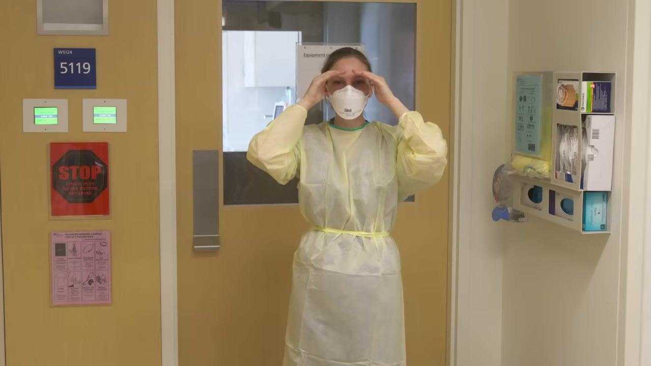 Healthcare worker in full gown and N95 respirator performing a seal check by placing hands around the mask.