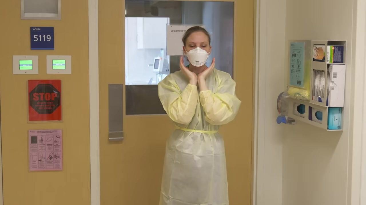 Healthcare worker in full PPE (gown and N95) preparing to don a face shield, hands positioned near the sides of the head.