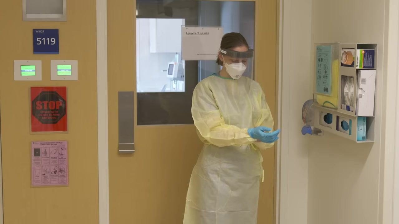 Healthcare worker in full PPE (yellow gown, N95, face shield) donning blue gloves in front of a patient room door. PPE supply station and signage visible.
