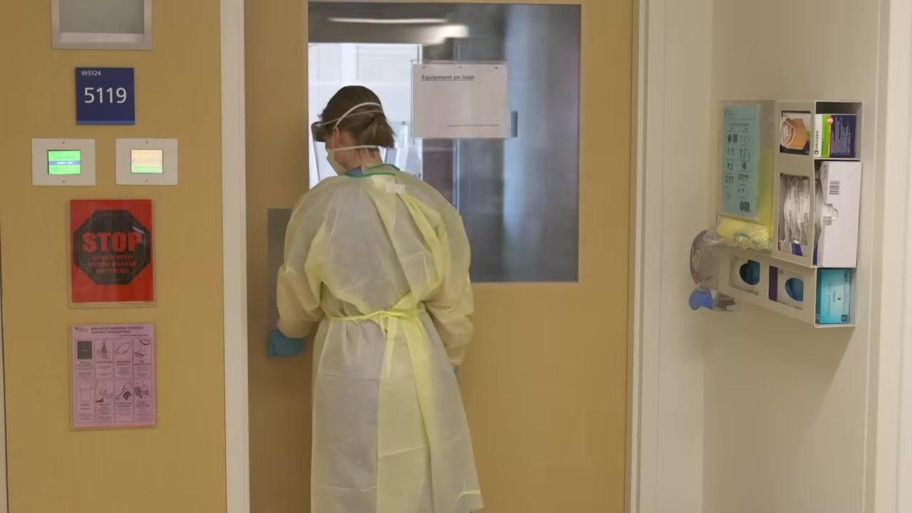 Healthcare worker in full PPE facing the door, reaching to open or close it. Room signage and PPE station visible.