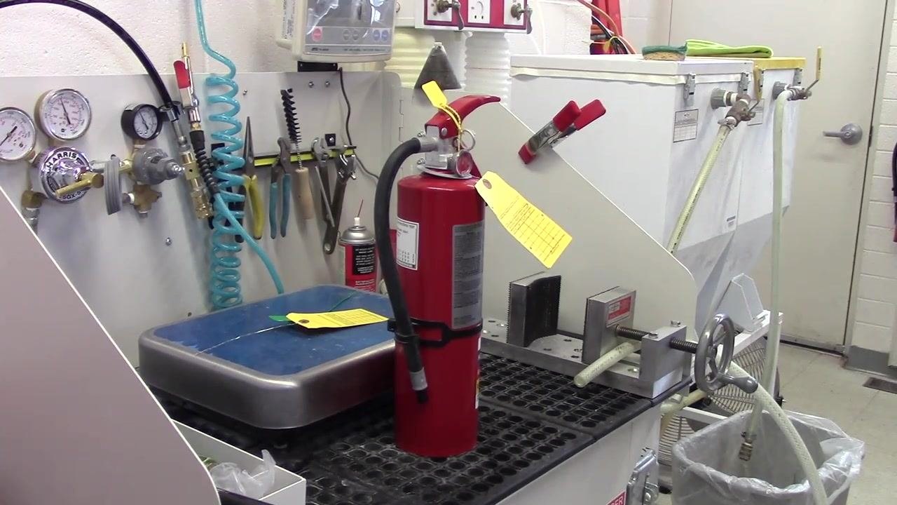 A fire extinguisher placed upright on a workbench in a maintenance area, surrounded by tools, gauges, hoses, and inspection tags.