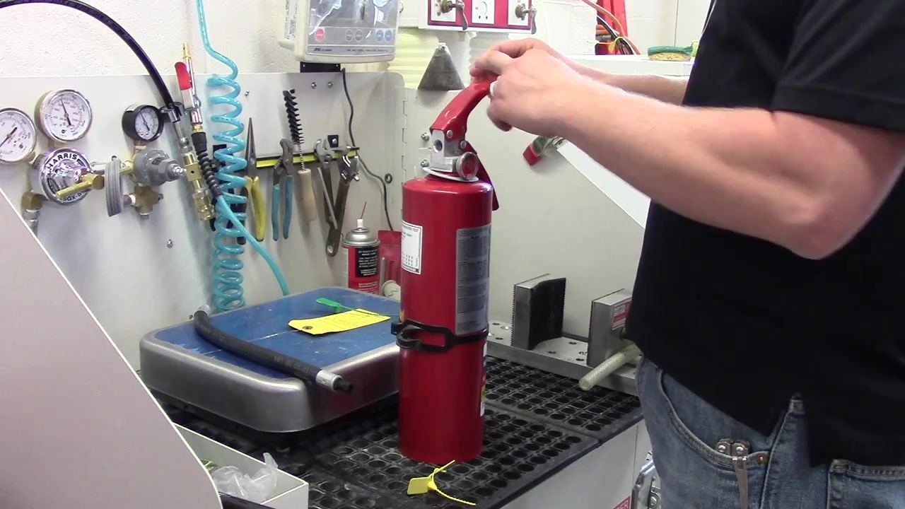 A person reinserting the safety pin through the handle and base of the fire extinguisher, with the extinguisher upright on a workbench. Gauges, tools, and hoses are visible in the background.