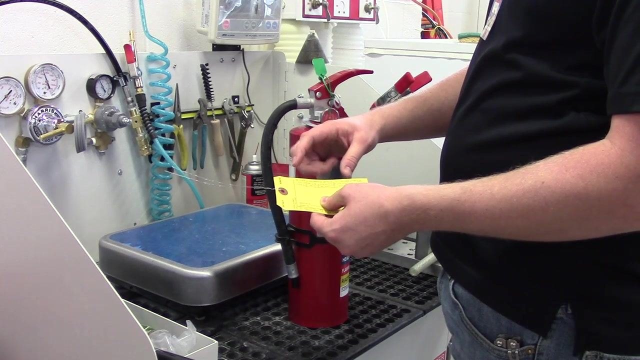 A person holding a yellow inspection tag next to a fire extinguisher, preparing to punch the tag. The workbench has various tools, gauges, and hoses visible.
