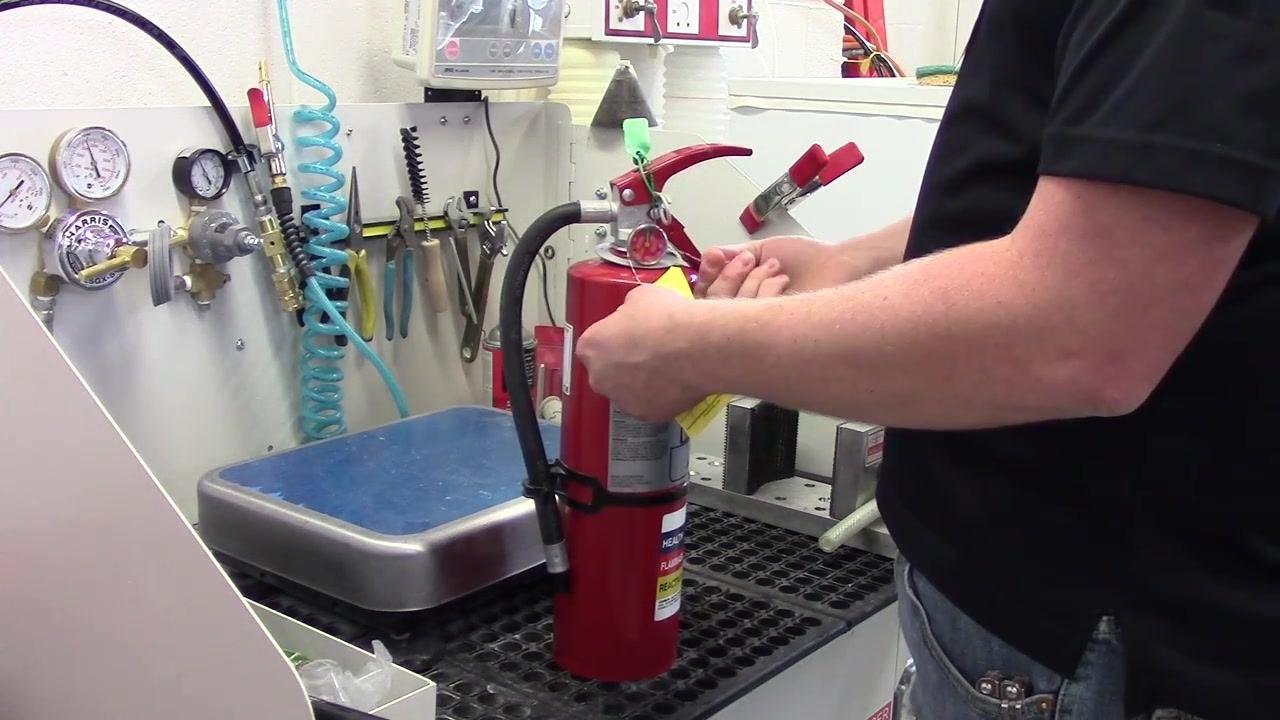 A person looping and securing the inspection tag wire around the pressure gauge of the fire extinguisher, pinching and wrapping it to hold the tag in place.