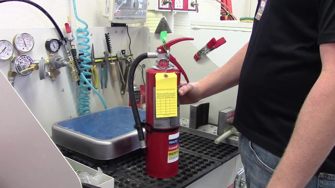 A completed fire extinguisher with a properly attached and positioned yellow inspection tag, ready for service. The workbench and tools are visible in the background.