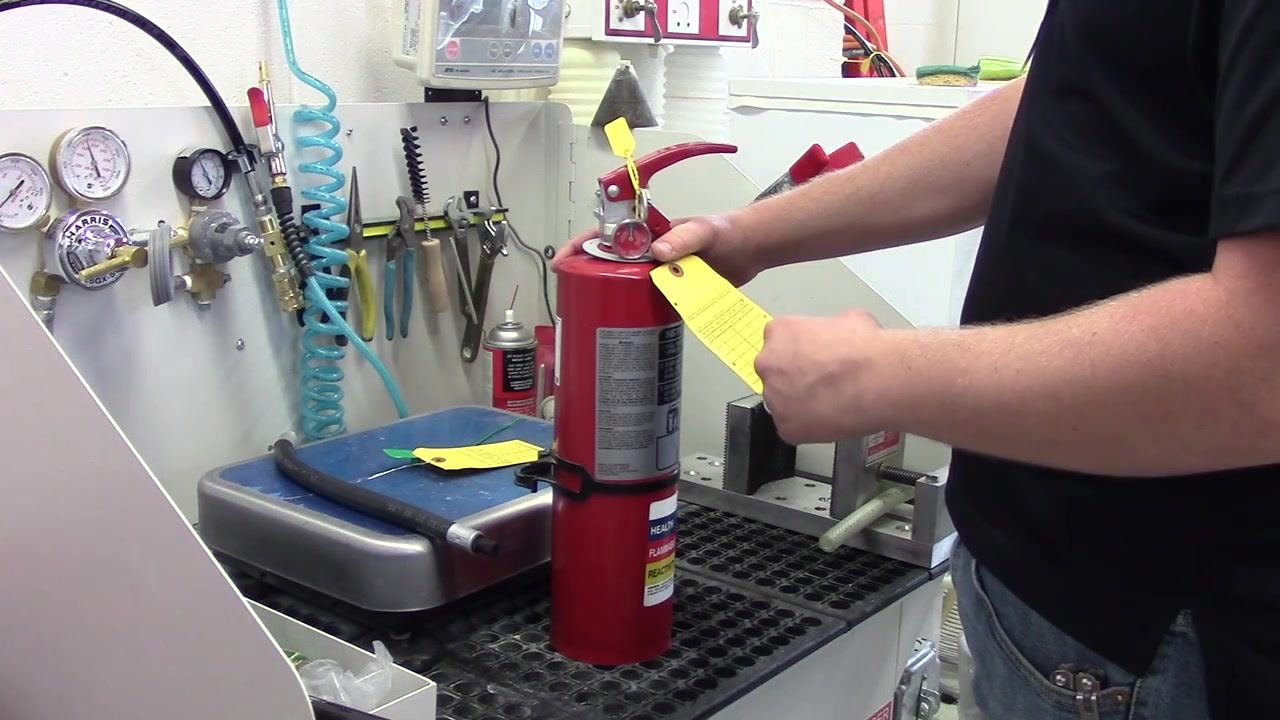 A person removing the yellow inspection tag from the fire extinguisher, with the extinguisher upright on a workbench. Gauges, tools, and hoses are visible in the background.