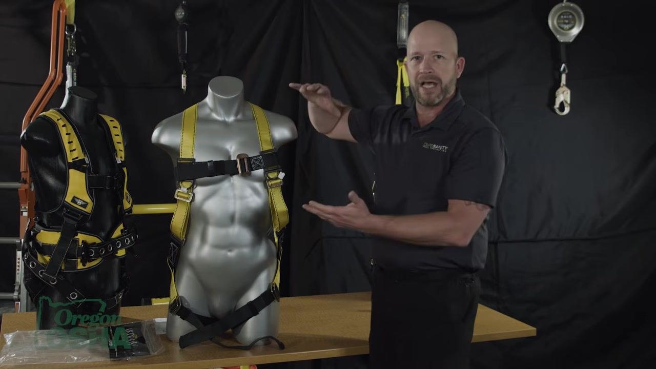 Gesturing with both hands at different heights next to a mannequin, explaining OSHA force requirements, with the mannequin facing forward showing the chest strap and harness configuration