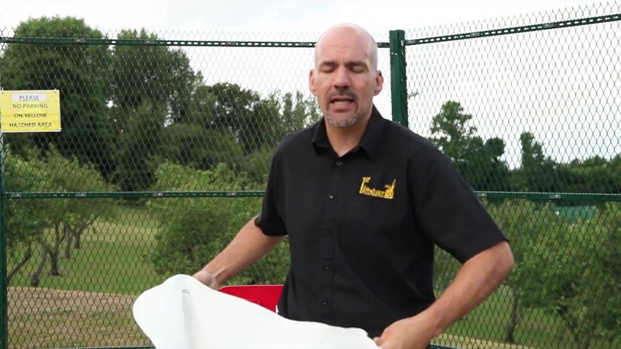 Person pointing to the location and mounting of a fire blanket on a red fire safety station, with a fence and greenery visible outdoors in the background.