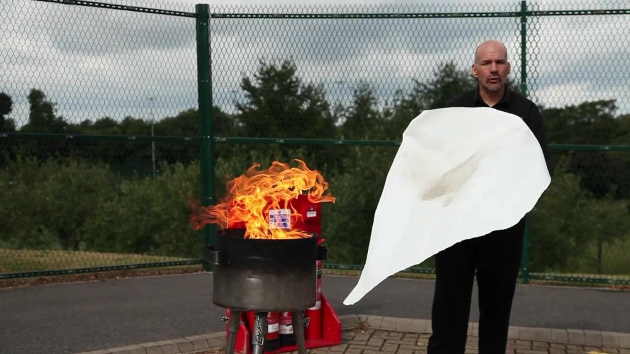 Person standing in front of a controlled fire, holding a fire blanket at the correct height and distance, ready to cover the flames.