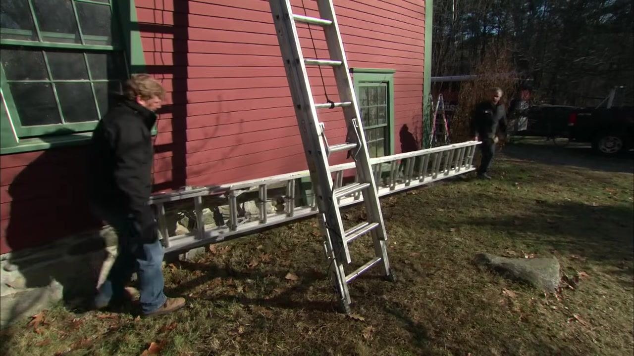 Two people carry a long extension ladder horizontally, positioning it against a red building with green trim.