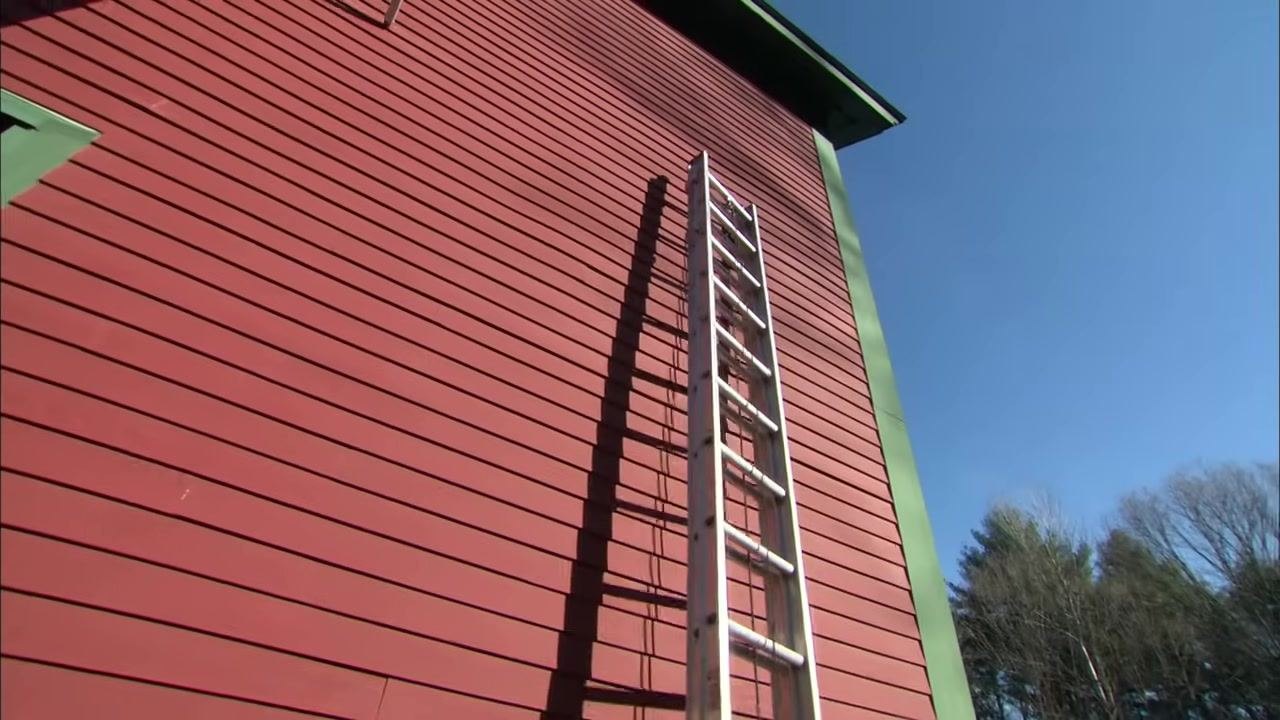 Two people place the base of an extension ladder against the foundation of a red building in preparation for raising it.
