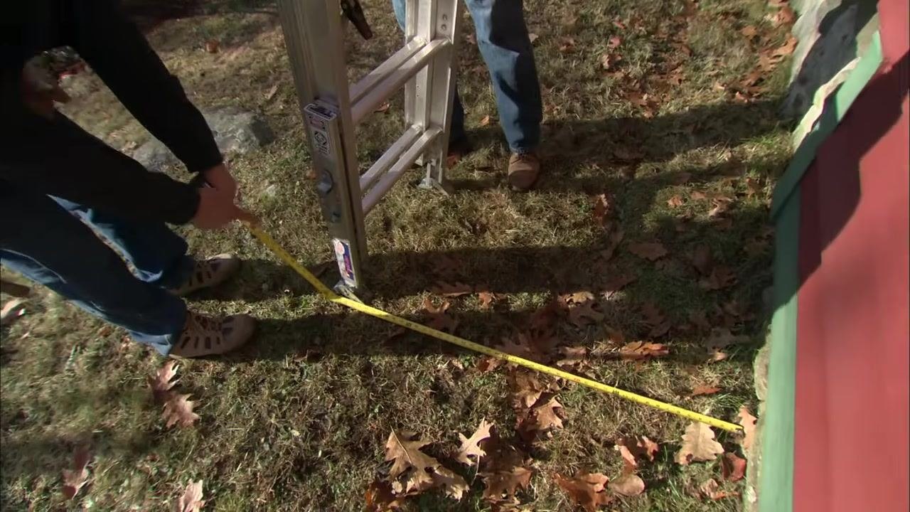 A close-up of a tape measure checking the distance from the base of the ladder to the building, with two people's legs and feet visible.
