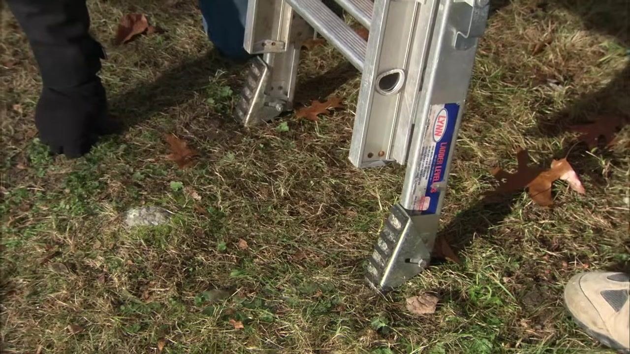 Two people inspect and adjust the feet of an aluminum extension ladder on a grassy surface, with the ladder's adjustment mechanisms visible.
