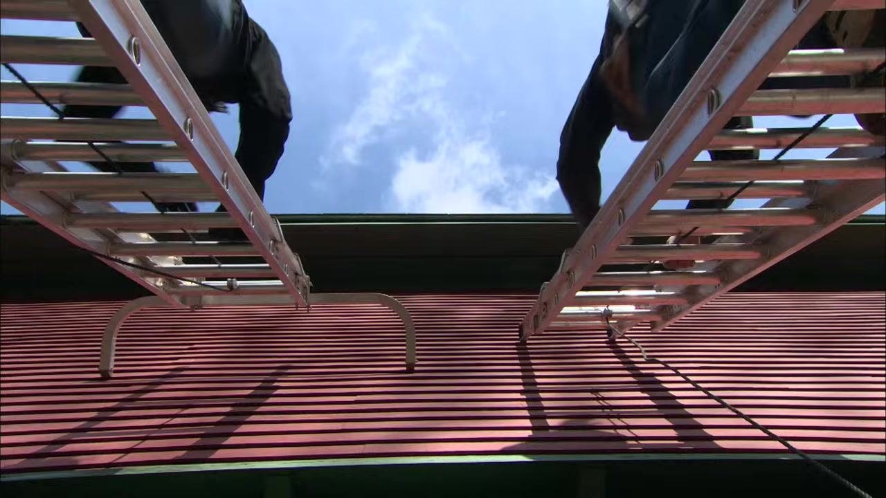 Two ladders with stabilizers attached at the top rest against the edge of a red building's roof, viewed from below as two people climb.