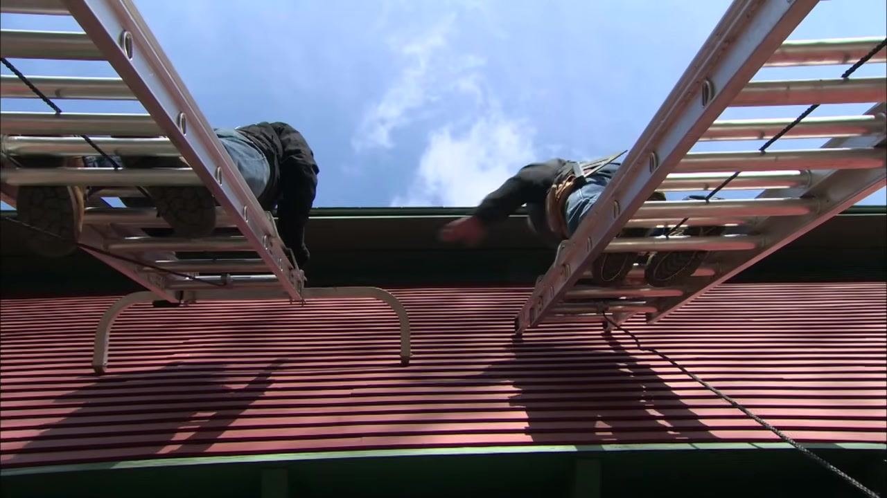 Two extension ladders with stabilizers attached at the top are set against a red building with green roof trim, keeping them clear of the gutter as two people climb.