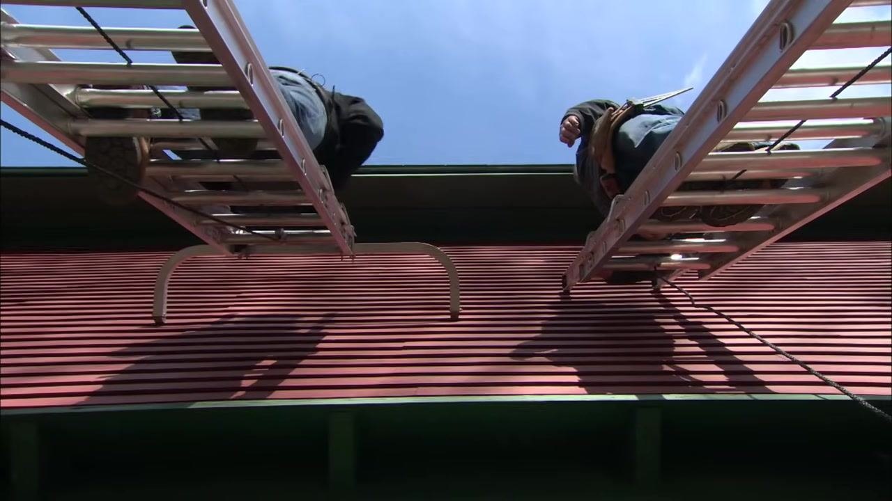 Extension ladders with stabilizers rest just below the roof overhang, keeping the ladders clear of the gutter as two people climb.