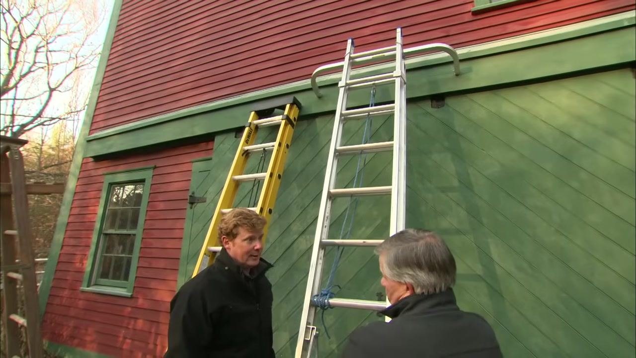 Two people stand outside a red and green building, pointing at a yellow fiberglass ladder and a silver aluminum ladder to compare the two options.