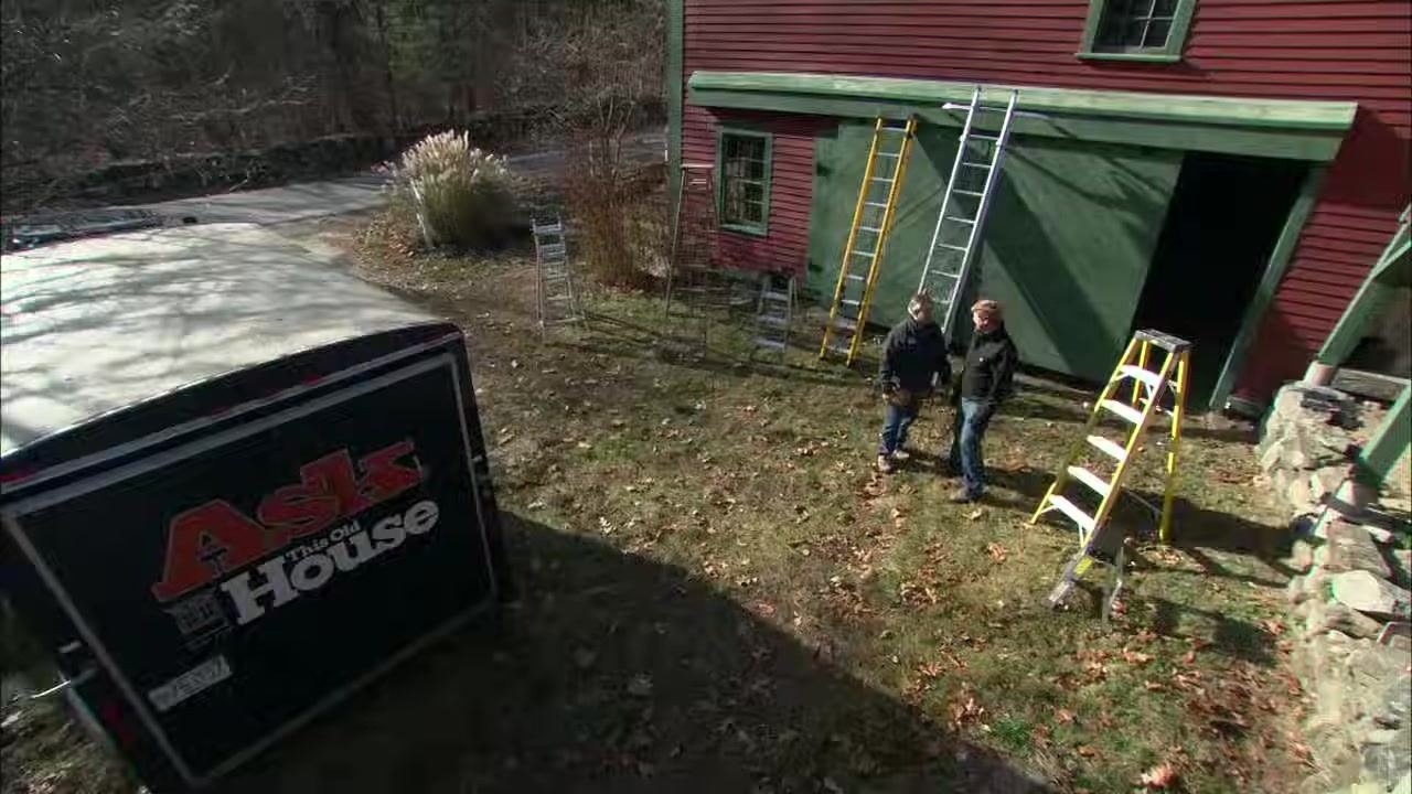 Two people stand in front of a red and green building discussing ladder safety, with multiple extension and step ladders set up around them in an outdoor yard.